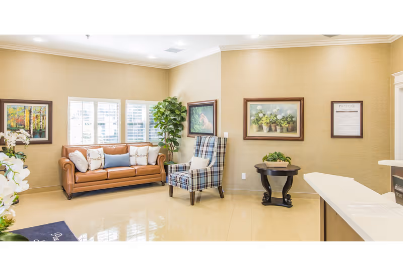 Bright reception seating area with a leather sofa, plaid armchair, framed artwork, and a small table in a senior living facility lobby.