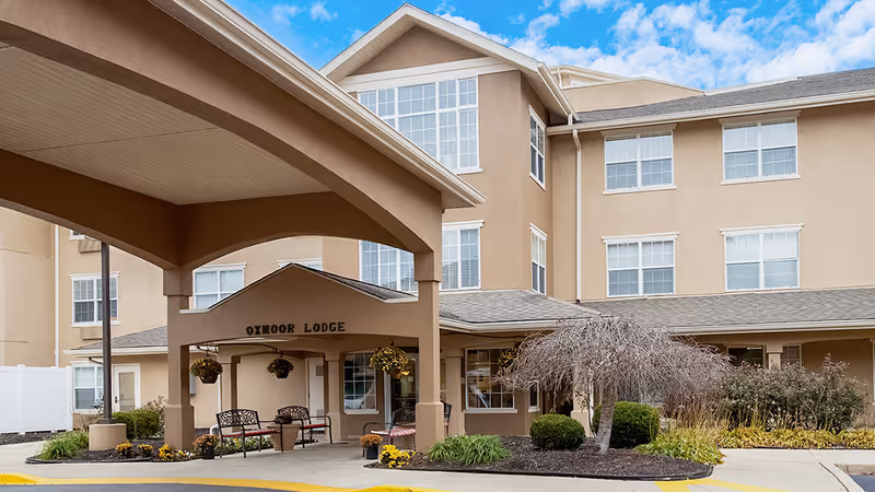 Exterior view of a senior living facility named Oxmoor Lodge, part of StoryPoint Louisville East, showing the building entrance with a covered drop-off area, benches, hanging flower baskets, and landscaping including bushes and a small tree.