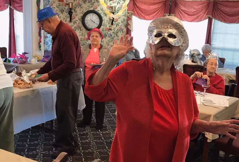 A group of elderly people in a decorated room, some wearing festive hats and masks. One woman in the foreground is wearing a silver mask and a red outfit, raising her hand. Other seniors are seated at tables with tablecloths, and one man is serving himself food from a buffet table. The room has floral wallpaper, a clock on the wall, and red curtains on the windows.