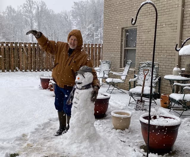 A man in a brown jacket stands beside a snowman on a snowy outdoor patio.