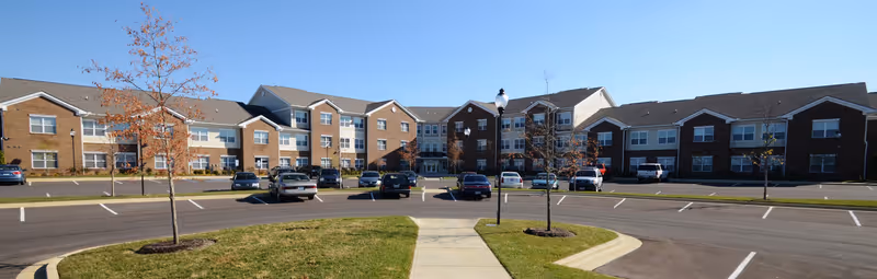 Wide exterior view of Hamburg Senior Residence, a large multi-story senior living building with brick and beige siding. There is a parking lot with several cars parked and a sidewalk leading to the main entrance. Small trees and grass areas are visible in the foreground under a clear blue sky.