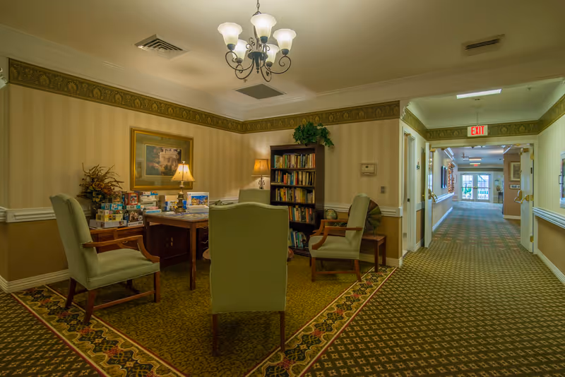 Carpeted common area with upholstered chairs around a table piled with board games, a bookshelf and lamps, and a long hallway leading to an exit.