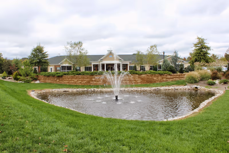 Exterior view of a senior living facility with a pond and a water fountain in the foreground, green grass surrounding the pond, and a building with a covered entrance and columns in the background under a cloudy sky.
