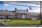 Three-story red brick building with a white columned entrance, an American flag flying in front, and a lawn with a sign.