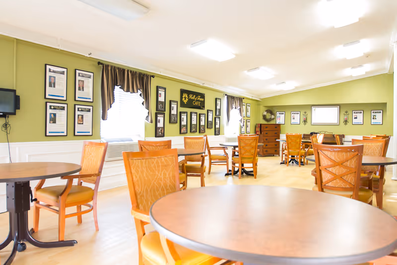 Bright communal dining room with round tables and wooden chairs, green walls decorated with framed photos and a small 'Café' sign.