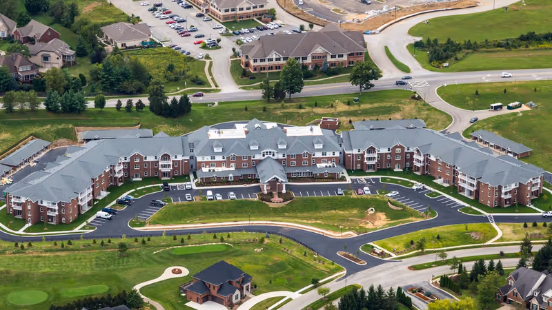 Aerial view of Village Crossing Retirement Community showing a large, U-shaped brick building with multiple floors and a gray roof, surrounded by parking lots, green lawns, and nearby roads. Additional residential buildings and trees are visible in the background.