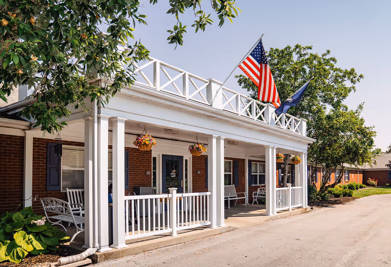 White-columned front porch of brick senior living facility with hanging planters and flags.