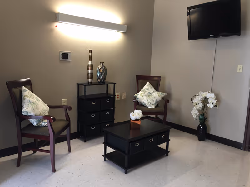 A small sitting area with two wooden armchairs with floral cushions, a black coffee table with a tissue box, a black shelving unit with decorative vases, a wall-mounted flat screen TV, and a vase with white flowers on the floor in the corner. The walls are painted beige and there is a long wall light fixture above the shelving unit.