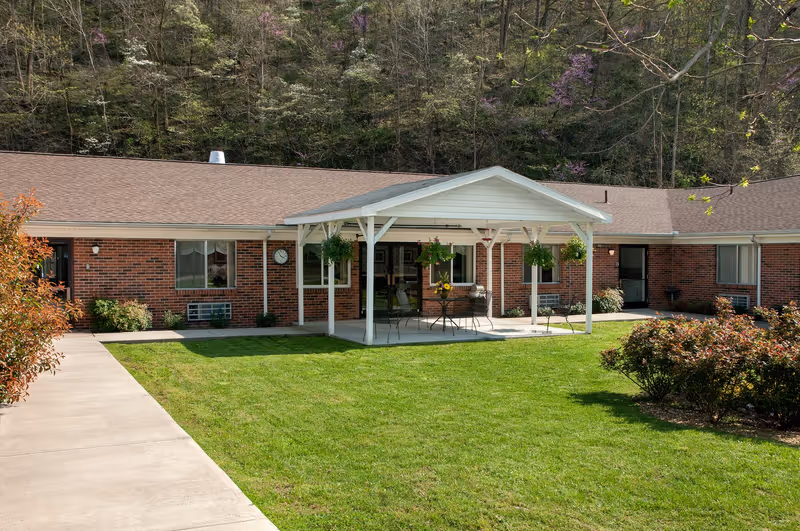 Brick senior living facility exterior with a covered patio, hanging plants, and a green lawn.