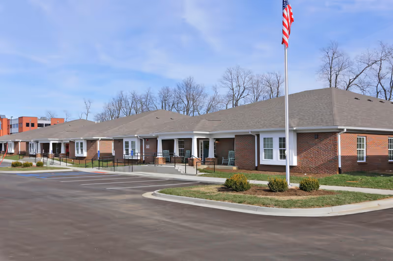 Exterior view of a single-story brick building with a gray roof, white window frames, and a covered porch area with rocking chairs. There is a flagpole with an American flag in front of the building, a paved parking lot with marked spaces, and some small bushes and grass landscaping. Leafless trees are visible in the background under a partly cloudy sky.