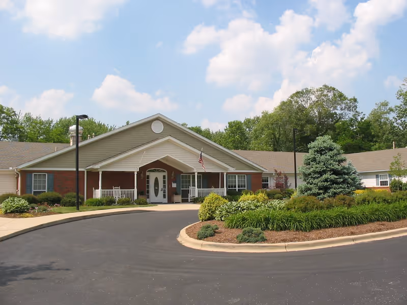 Front exterior view of a single-story senior living facility building with a covered entrance, white pillars, an American flag, and landscaped greenery including bushes and a small tree. The sky is partly cloudy.