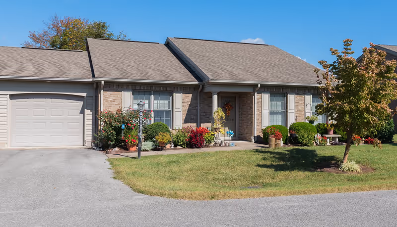 Single-story brick residential building with a garage on the left, a front porch with a door decorated with a wreath, and windows with shutters. The front yard has a well-maintained lawn, various bushes, flowers, and a small tree. The sky is clear and blue.