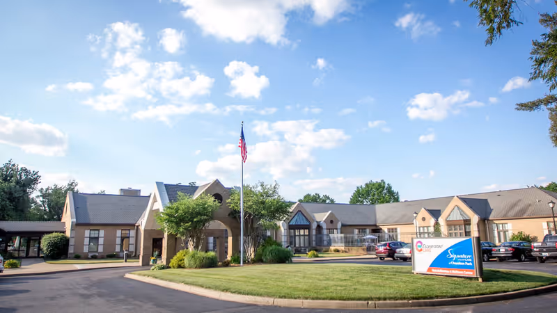 Exterior view of Signature HealthCARE of Cherokee Park building with a flagpole flying the American flag in front, surrounded by green grass and trees under a partly cloudy blue sky. Several cars are parked near the entrance.