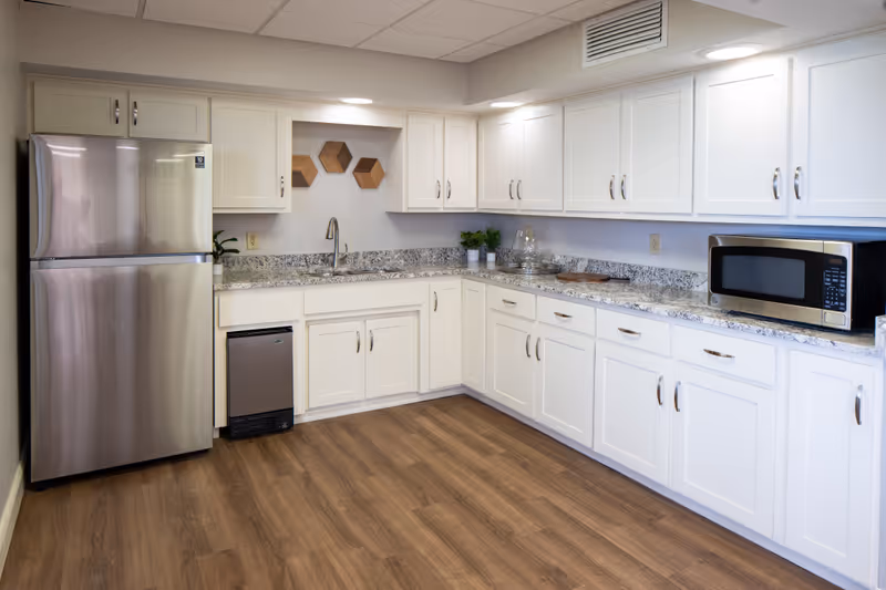 A modern kitchen with white cabinets, granite countertops, a stainless steel refrigerator, a small under-counter refrigerator, a microwave, and a sink. The floor is wood, and there are decorative hexagonal wooden shelves on the wall above the sink.