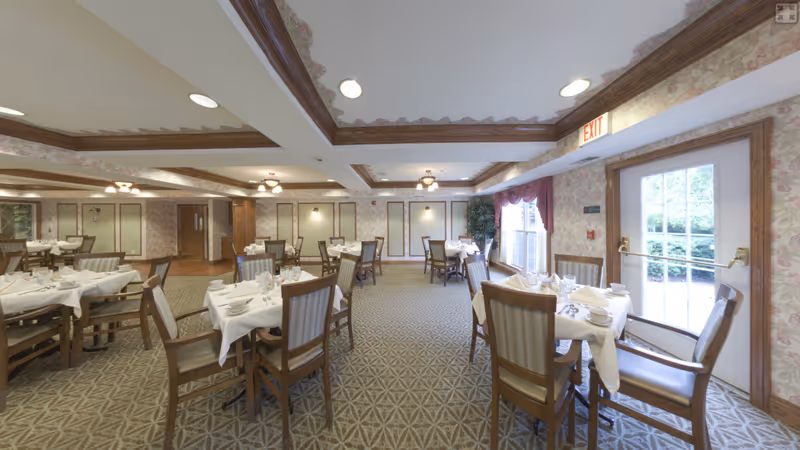 A dining room in a senior living facility with multiple tables set with white tablecloths, napkins, cups, and glasses. The room features patterned carpet, floral wallpaper, wooden trim, and large windows with curtains letting in natural light. There is an exit door on the right side of the image.
