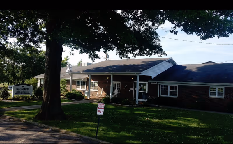 Exterior view of Springfield Nursing & Rehabilitation Center, a single-story brick building with a covered entrance, white rocking chairs on the porch, surrounded by green grass and trees. A sign near the entrance reads 'Springfield Nursing & Rehabilitation'.