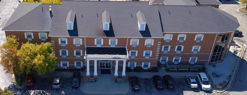 Aerial view of the three-story brick Village Care Center building with a columned entrance and parked cars in front.