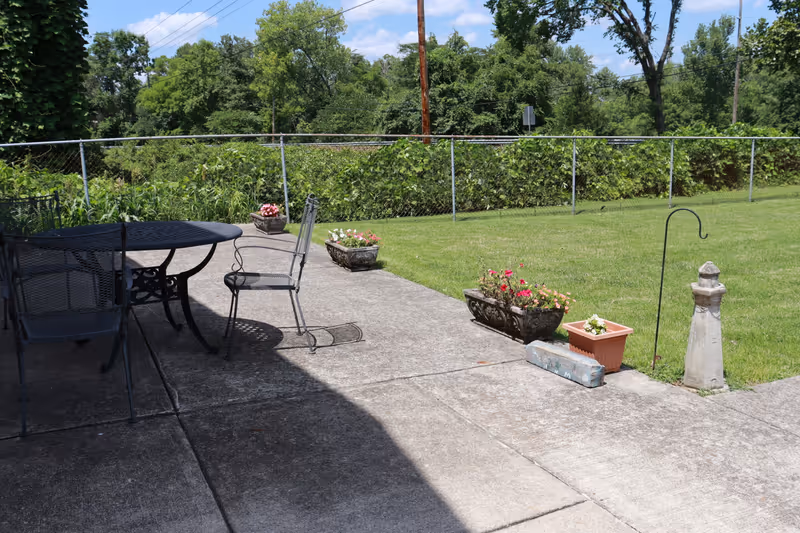 Outdoor patio area with metal table and chairs on a concrete surface, several flower pots with blooming flowers along the edge, a grassy lawn, and a chain-link fence with trees and blue sky in the background.