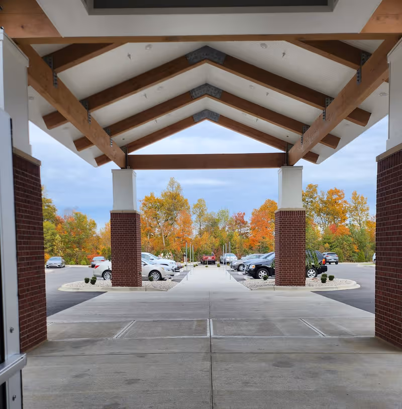 Covered entrance with wooden beams and brick columns over a driveway; autumn trees in background.