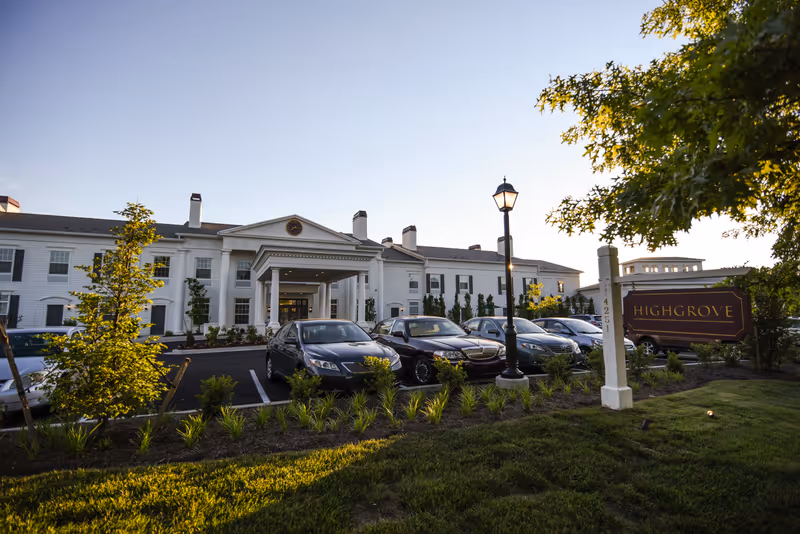 Exterior view of the Highgrove at Tates Creek senior living facility during sunset, showing a white two-story building with a covered entrance, several parked cars, a street lamp, landscaped greenery, and a sign with the facility name.