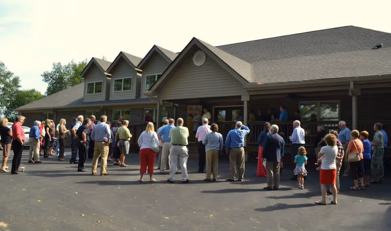 A group of people standing outside a large building with a porch, listening to a speaker. The building has multiple windows and a sloped roof, and the scene is set on a paved area with trees in the background.