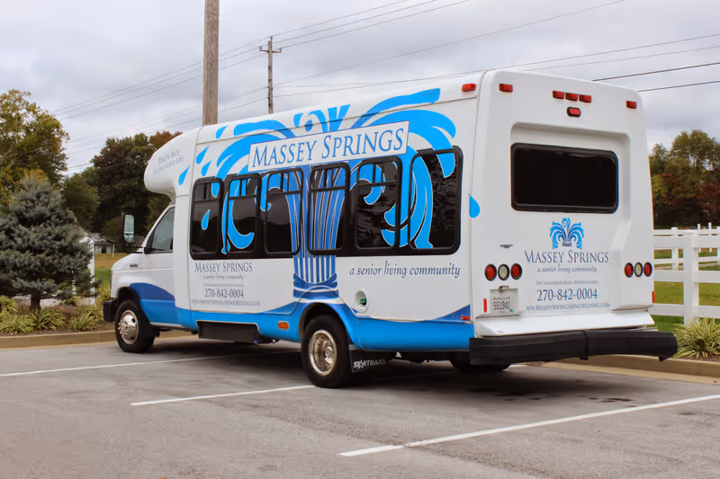 A white and blue shuttle bus parked in a parking lot with the branding 'Massey Springs, a senior living community'. The bus features a large blue water fountain graphic and contact information including a phone number and website. Trees and a white fence are visible in the background under a cloudy sky.