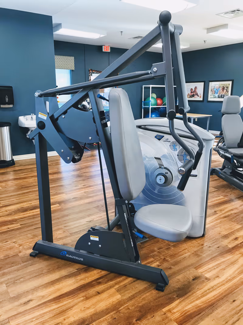 A fitness machine with a padded seat and backrest in a room with wooden flooring and blue walls. In the background, there are exercise balls on a rack, framed pictures on the wall, and another exercise machine.
