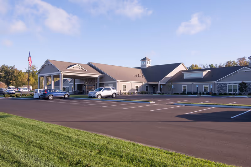 Exterior view of The Lantern at Morning Pointe Alzheimer's Center of Excellence, Russell, showing a large single-story building with a covered entrance, several parked cars, an American flag, and a spacious parking lot under a partly cloudy sky.