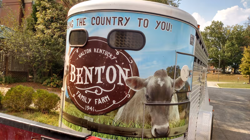 Rear of a large bus painted with Benton Farm mural outdoors beside trees.