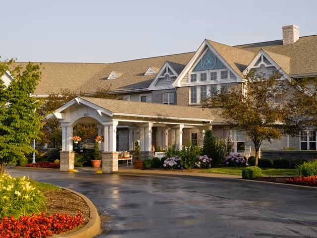 Front exterior view of Vitality Living Springdale facility showing a large building with a covered entrance, landscaped garden beds with flowers and shrubs, and a paved driveway.