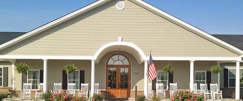 Front exterior view of a senior living facility with a beige facade, white columns, a central arched doorway with glass panels, several white rocking chairs on the porch, hanging green plants, and an American flag displayed near the entrance.