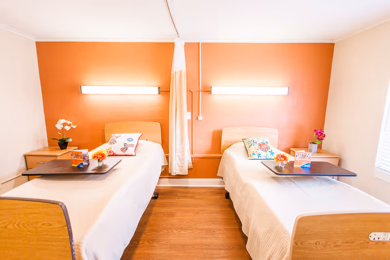 A bright and clean nursing facility bedroom with two single beds separated by a curtain. Each bed has a wooden headboard, white bedding, a colorful pillow, and a small overbed table with a flower and a card. There are two wooden nightstands with potted plants on each side of the beds. The back wall is painted orange with two horizontal wall lights above each bed. The floor is wooden, and there is a window with blinds on the right side.