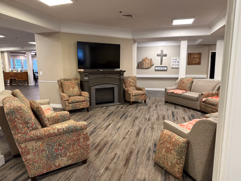 A cozy living room area in a senior living facility with patterned armchairs and beige sofas arranged around a dark fireplace with a mounted flat-screen TV above it. The room has wood-look flooring and neutral-colored walls decorated with inspirational wall art including a cross. The space is well-lit with ceiling lights and has an open view into an adjacent room with large windows.