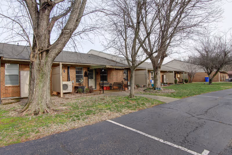 Row of brick senior living cottages with porches, trees, and a paved parking lot.