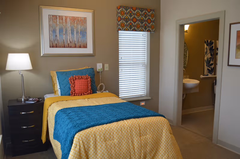 A neatly decorated bedroom with colorful bedding, a window, and an adjacent bathroom.