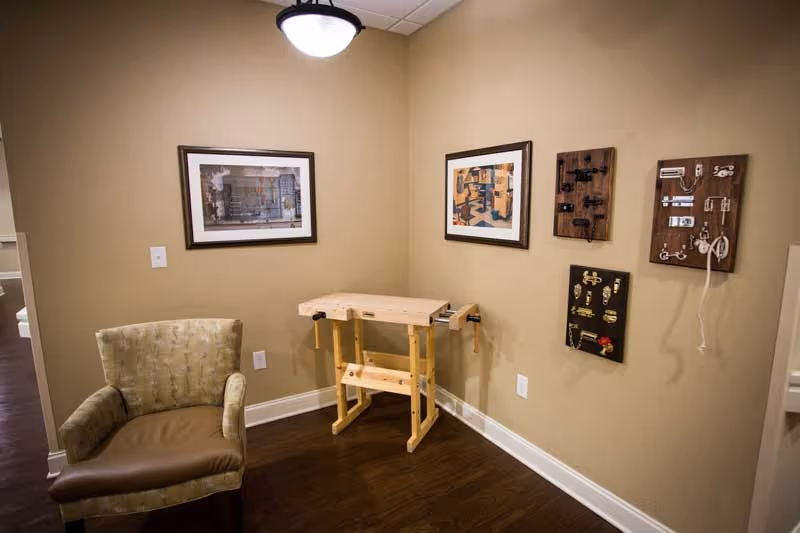 Corner of a memory-care activity room with an upholstered armchair, a small wooden workbench, framed artwork, and several tactile activity boards on the wall.