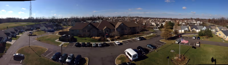 Aerial panoramic view of Liberty Ridge Senior Living facility showing multiple buildings with brown roofs, parking lots with several cars, a flagpole with an American flag, and surrounding residential neighborhood under a clear blue sky.