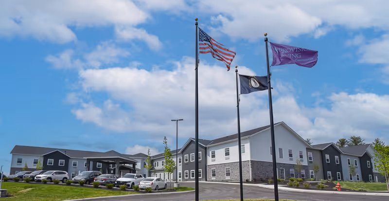 Exterior view of a senior living facility building with a parking lot in front. Three flagpoles display the American flag, a state flag, and a purple Provision Living flag. The sky is partly cloudy.