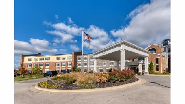 Exterior view of Cedarhurst of Beaumont senior living facility showing a large multi-story building with a covered entrance, landscaped roundabout with flowers and shrubs, an American flag on a flagpole, and a partly cloudy blue sky.