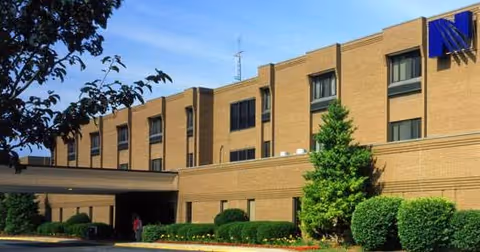 Exterior front of a multi-story tan brick senior living building with a covered entrance and landscaped shrubs.