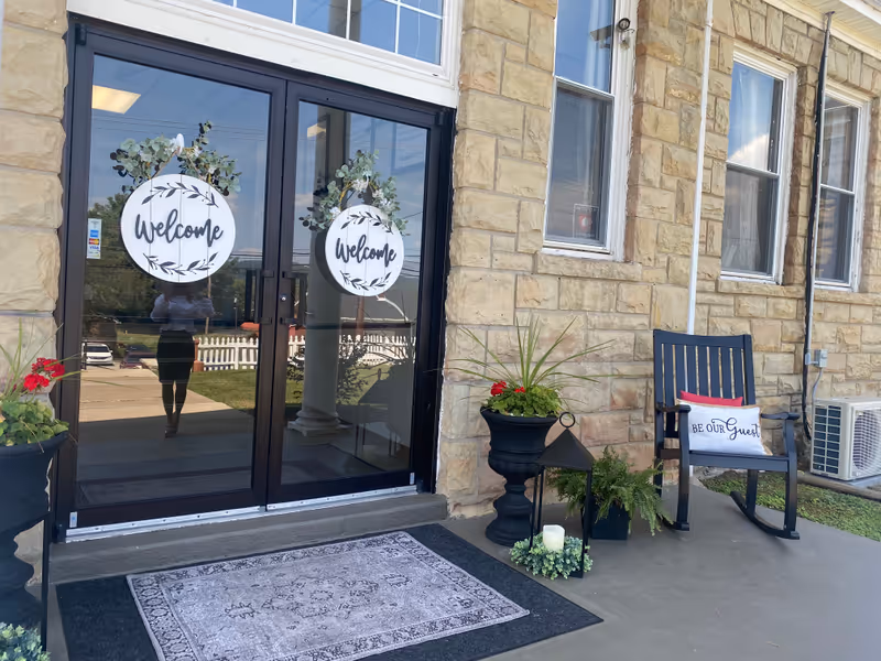 Stone building exterior with glass doors, welcome signs, a rocking chair, and potted plants.