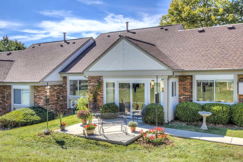 Front exterior of a brick senior living unit with a small patio, chairs, potted plants, and a manicured lawn.