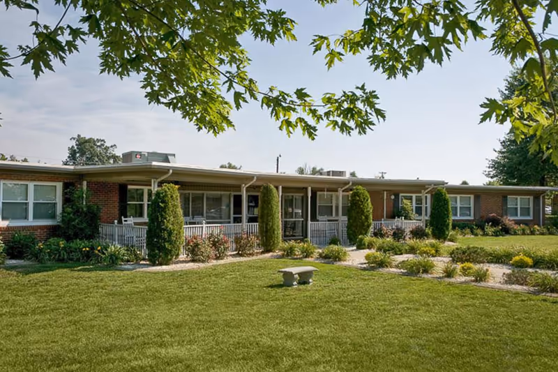 Single-story brick building with a covered porch, surrounded by well-maintained green lawn, shrubs, and trees under a clear sky.