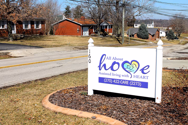 A white sign with purple and blue text reading 'All About hope Assisted living with HEART' and a phone number (270) 422-CARE (2273) is placed on a mulched area next to a road. Behind the sign, there are several single-story brick buildings and leafless trees under a clear sky.