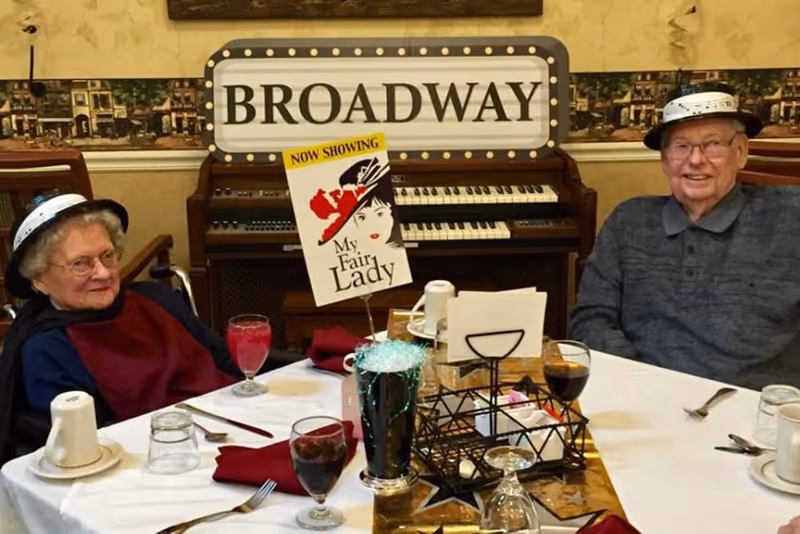 Two elderly people sitting at a dining table set with glasses, cups, and utensils. They are wearing hats and smiling. Behind them is a piano with a sign that reads 'BROADWAY' and a poster for the musical 'My Fair Lady.'