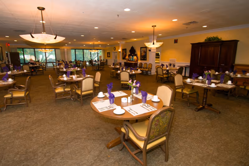 Spacious dining room with round tables set with place settings, chairs, and purple napkins in a senior living facility.