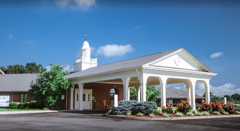 Front entrance of a single-story brick nursing facility with a white porte-cochere, cupola, and landscaped flowerbeds.