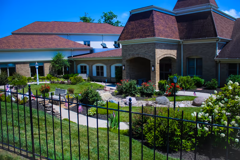 Exterior view of Carmel Manor senior living facility showing a well-maintained garden with green grass, bushes, flowers, and small American flags along a paved walkway. The building has a brick facade with a brown shingled roof and arched entryways. A black metal fence is visible in the foreground under a clear blue sky.
