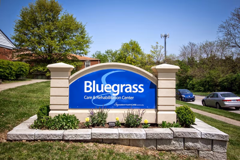 Outdoor view of a large blue sign for Bluegrass Care & Rehabilitation Center, set in a landscaped stone planter with flowers and greenery, with trees, a sidewalk, and parked cars in the background under a clear blue sky.