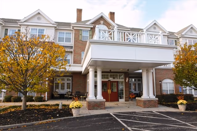 Front exterior view of a multi-story senior living facility building with brick facade, white columns supporting a covered entrance, red double doors, and autumn trees with yellow leaves on either side of the entrance.